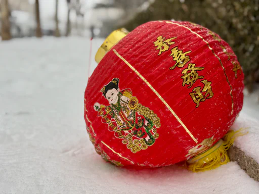 A red Chinese lantern with gold lettering and a colourful figure, resting on snow.